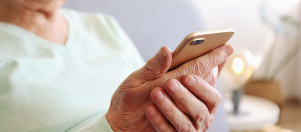 Elderly woman holding blank screen cell phone gadget in hands. Old lady with wrinkled skin trying to vigure out touchscreen smartphone. Background, close up, overhead, top view, copy space.