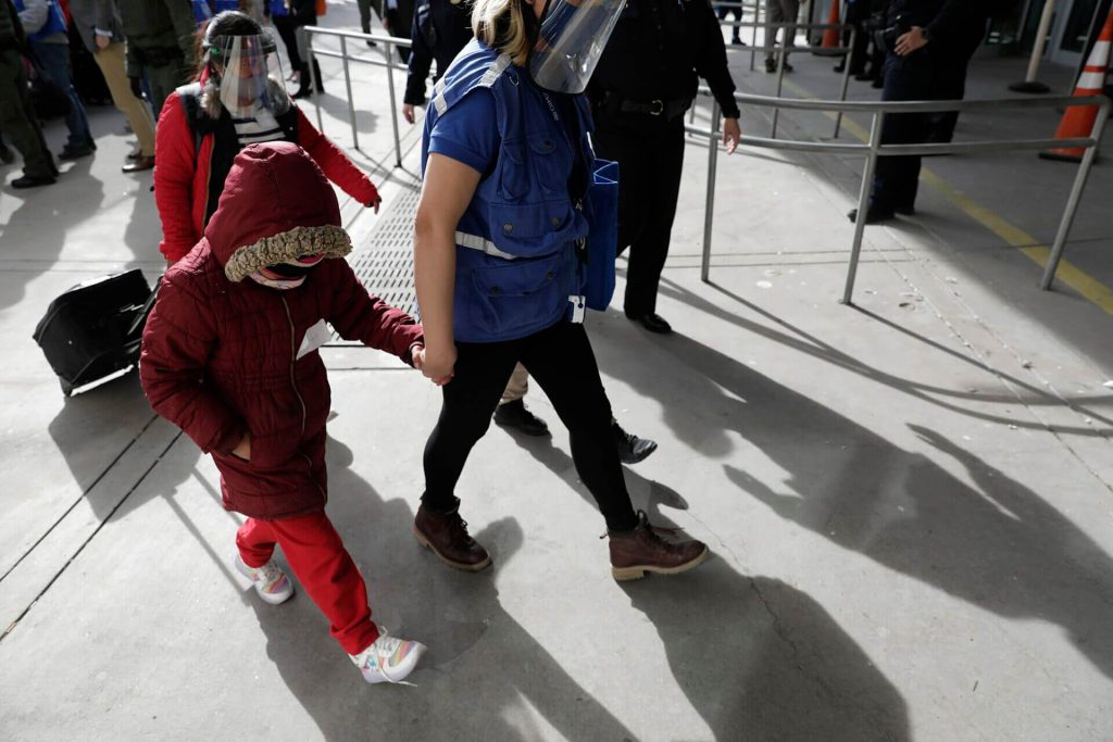 Este artículo habla sobre la política migratoria "Quédate en México". La foto muestra una oficial de frontera escoltando a un grupo de personas que solicitan asilo en uno de los puntos de entrada a los Estados Unidos.